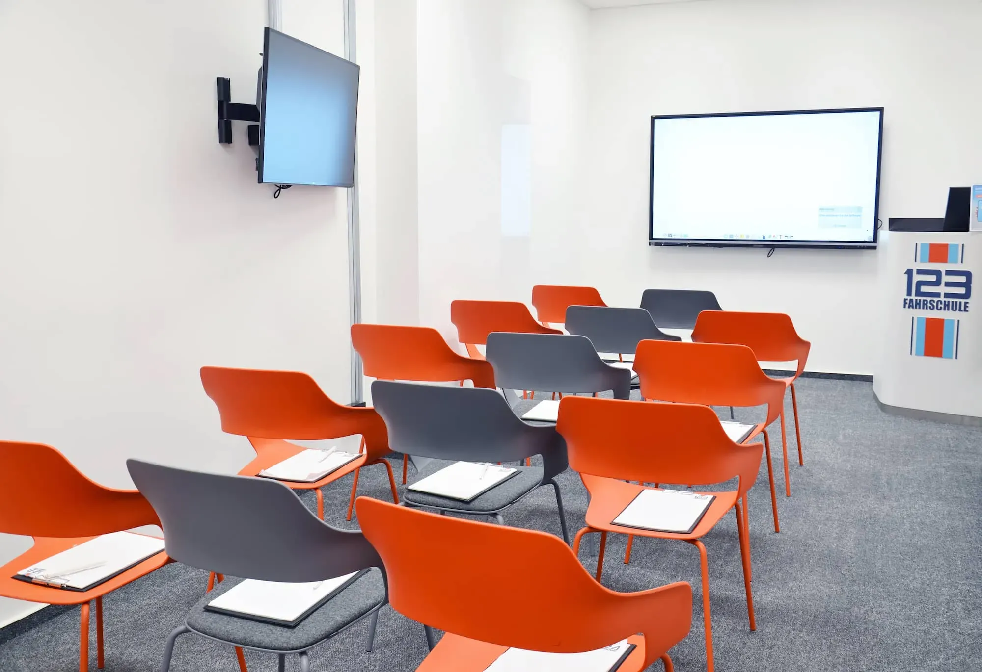 Theorieraum einer Filiale der 123fahrschule mit orangefarbenen und grauen Stühlen und zwei Bildschirmen